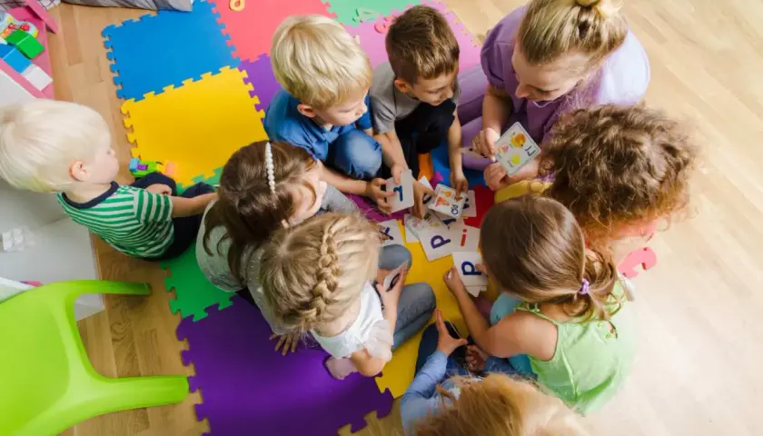 Dzieci i dorosła siedzą na kolorowych puzzlach, grając w karty z literami.