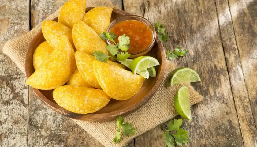 Empanadas colombianas doradas en plato de madera con salsa, limones y cilantro sobre mesa rústica.