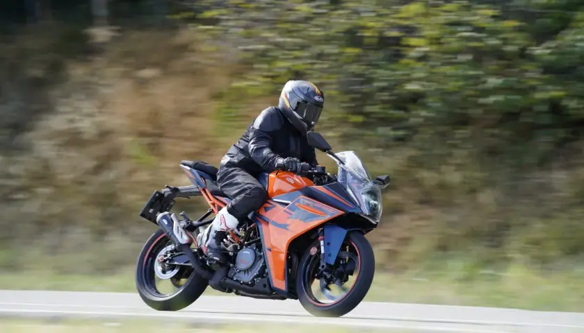 Motociclista con casco y traje negro conduce una KTM RC 390 CV naranja y azul por una carretera con árboles borrosos al fondo.