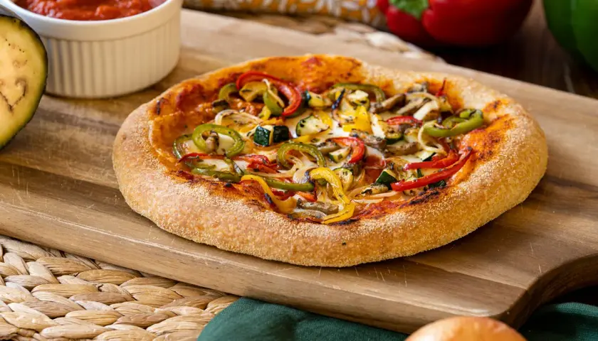 Pizza vegana con pimientos, calabacín y champiñones sobre tabla de madera. Salsa de tomate y berenjena al lado.