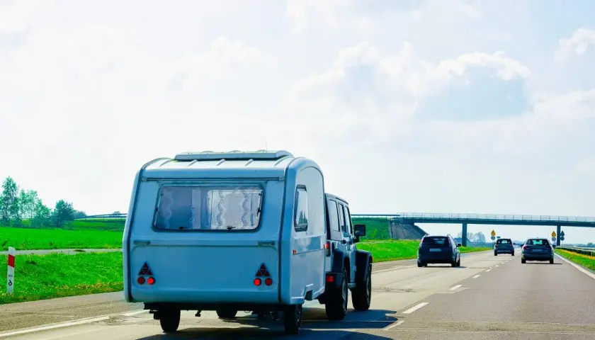 Samochód terenowy ciągnie dmc przyczepy kempingowej po autostradzie. W tle zielone pola i most.