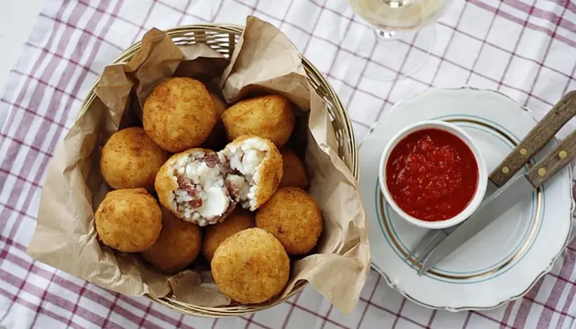 Koszyk pełen złotych arancini, jedno przekrojone ukazuje ryż i ser. Obok miseczka z sosem pomidorowym. Idealne na przekąskę, gdy masz ochotę na arancini przepis.