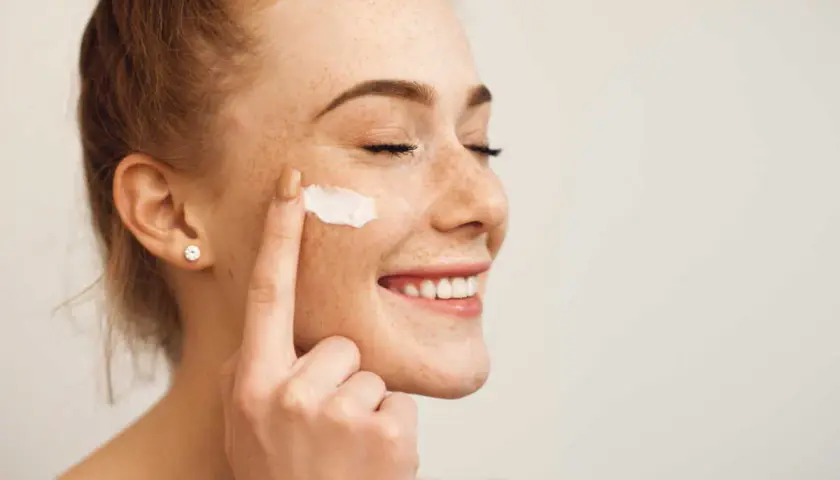 Une jeune femme rousse souriante, les yeux fermés, applique de la crème sur sa joue. Elle montre comment appliquer crème visage pour une peau éclatante.