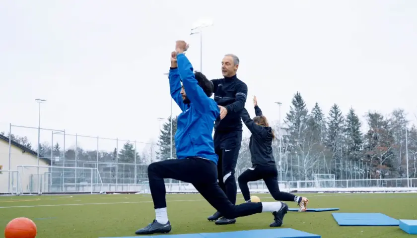 Athletik Training Fußball: Trainer korrigiert Ausfallschritt bei Spieler auf Kunstrasen. Eine Spielerin macht mit.
