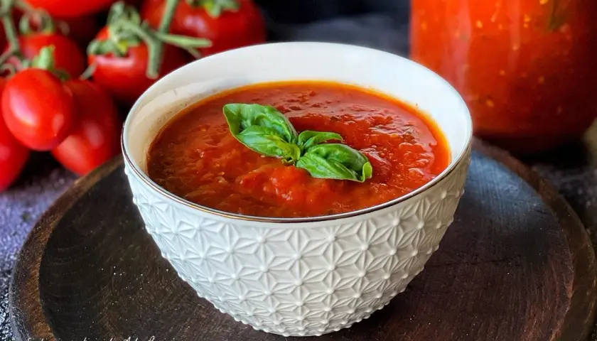 Una ciotola di salsa di pomodoro fatta in casa, guarnita con basilico fresco, con pomodori maturi sullo sfondo.