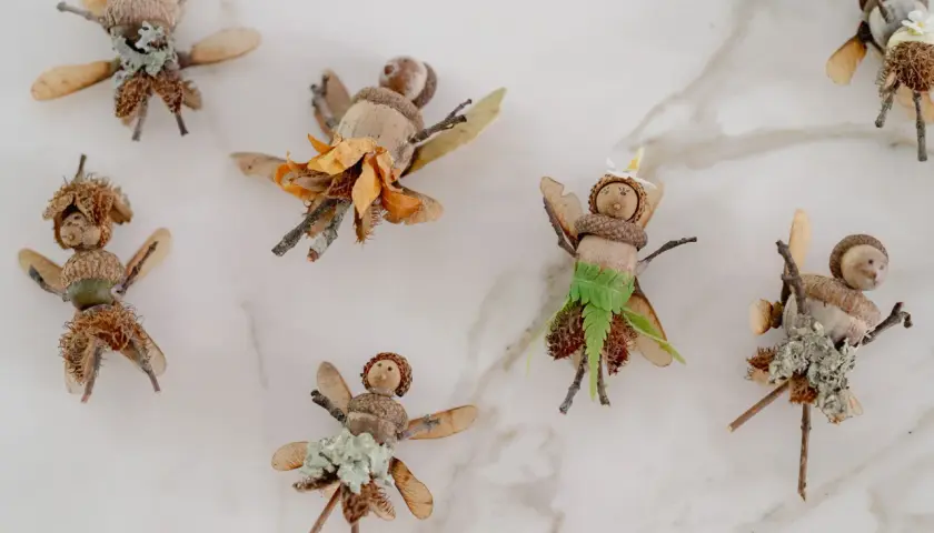 Figuras de hadas hechas con bellotas, ramas y hojas sobre fondo blanco.