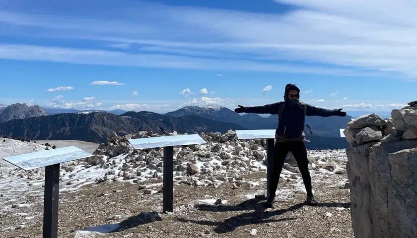 Persona amb els braços oberts al cim del Pedra dels Quatre Batlles, amb paisatge muntanyós i cel blau.