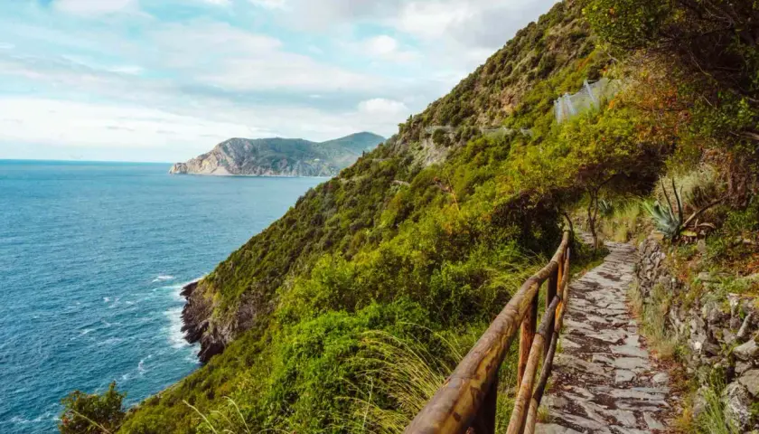 A stone path winds along the coast for Cinque Terre hiking, with lush green hills and the blue sea stretching to the horizon.