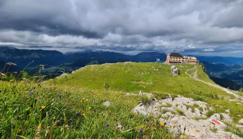 Watzmannhaus Wanderung: Ein Berggasthof thront auf einem grünen Kamm, umgeben von blühenden Almwiesen und dramatischen Wolken.