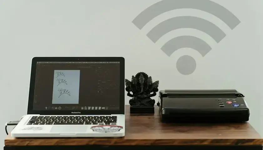 Laptop, drukarka i figurka Ganesha na biurku. Ikona Wi-Fi na ścianie sugeruje, jak połączyć drukarkę z wifi bezprzewodowo.