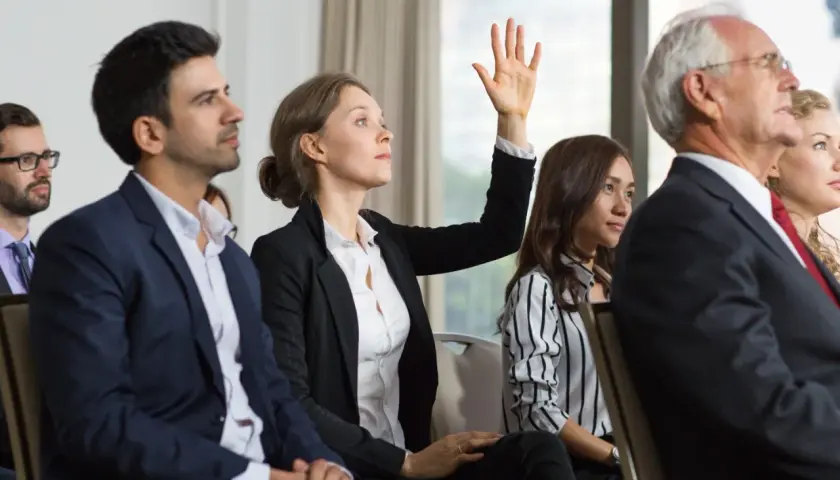 Une femme lève la main lors d'une réunion, posant une question sur le délai entre 2 ag de copropriété.