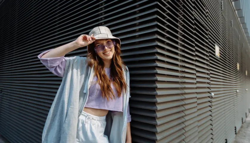 Chica vsco con sombrero y gafas de sol moradas, sonriendo y apoyada en una pared de lamas grises.