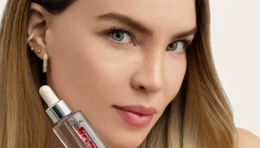 Mujer con cabello rubio y joyas brillantes sostiene un frasco de sérum, parte de su rutina de cuidado de la piel.