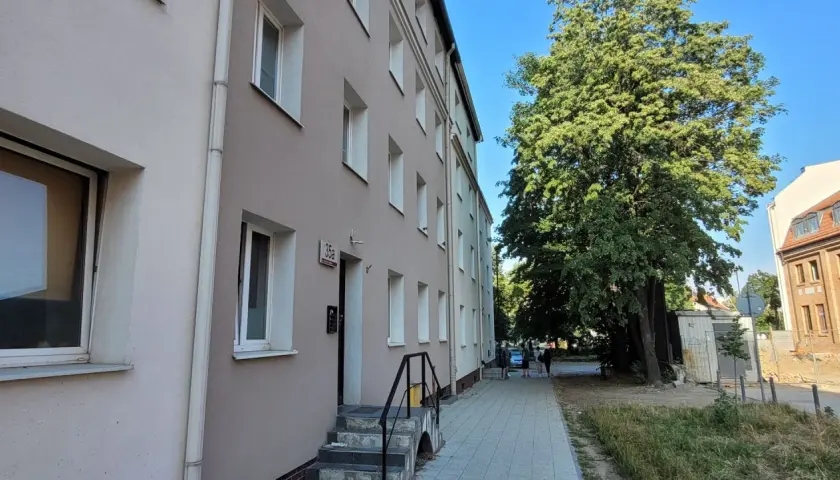 Budynek mieszkalny z oknami i drzwiami, obok drzewa i chodnik. To miejsce może być związane z art. 18 ustawy o ochronie praw lokatorów.