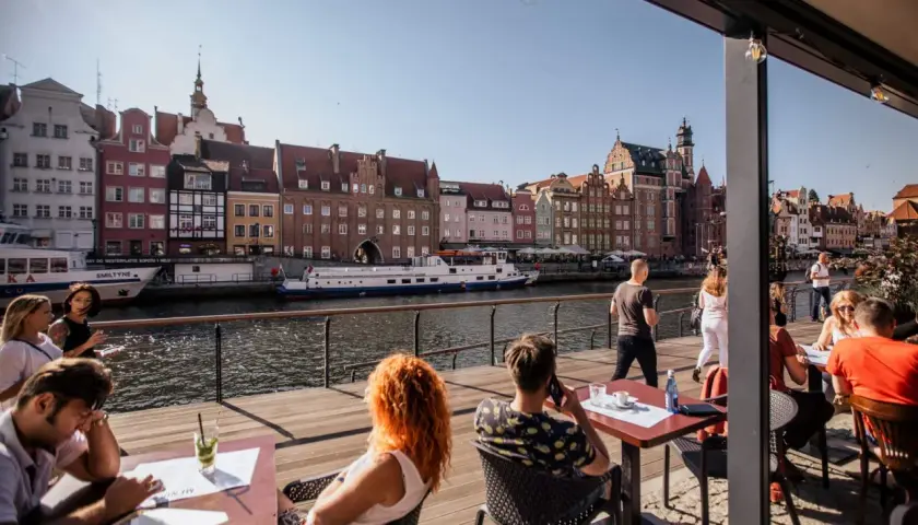 Gdańsk restauracje z widokiem na Motławę. Ludzie siedzą przy stolikach na drewnianym tarasie, podziwiając zabytkową zabudowę i pływające statki.