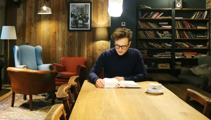 Mężczyzna po francusku, zanurzony w lekturze przy kawie, w przytulnej kawiarni z drewnianymi ścianami i regałami pełnymi książek.