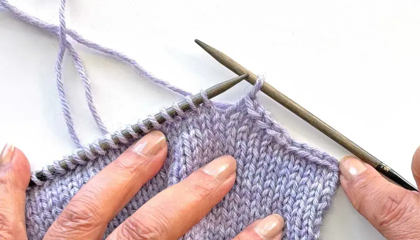 Close-up of hands sewing shoulder seams in knitting with lilac yarn and wooden needles.
