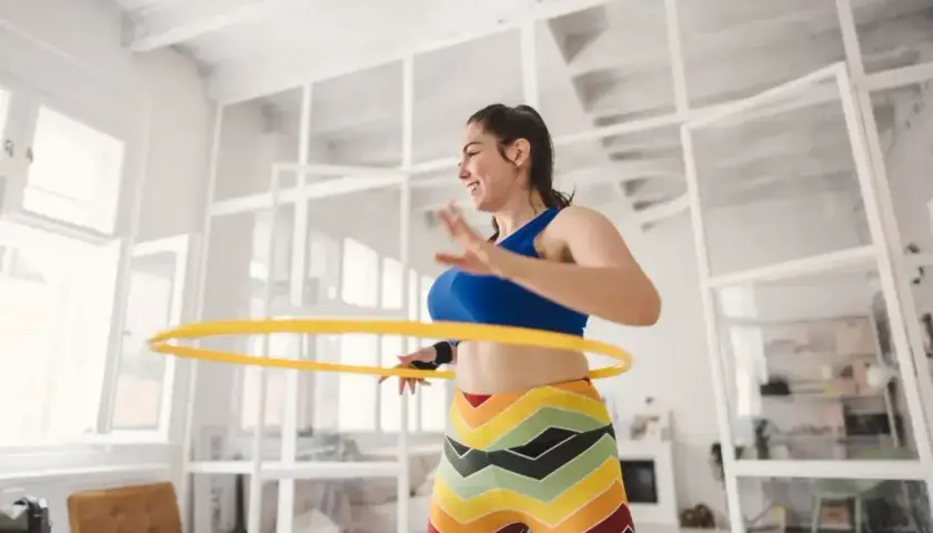 Uśmiechnięta kobieta kręci żółtą obręczą hula hop. Ćwiczenie to nie jest szkodliwe, a wręcz przeciwnie – świetnie wpływa na sylwetkę.