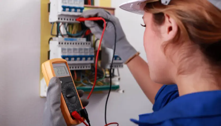 Elektryk w białym kasku wykonuje pomiary elektryczne przy skrzynce z bezpiecznikami, trzymając multimetr.