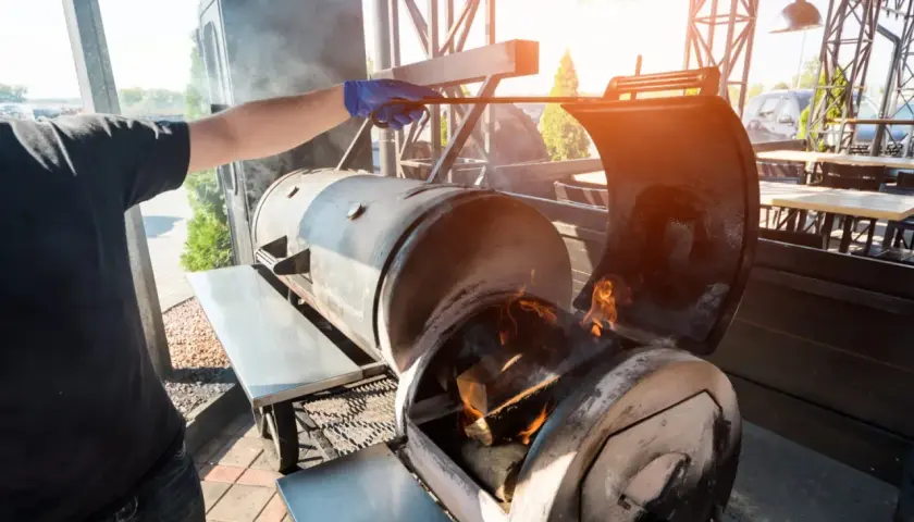Un homme ajuste le bois pour fumoir dans un grand barbecue. Des flammes dansent dans le foyer, promettant une cuisson savoureuse.