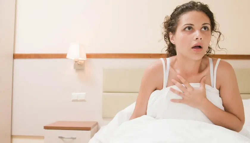 Mujer despierta en cama, con expresión de asombro y mano en el pecho, como si acabara de tener un sueño vívido: soñar que mi padre fallecido está vivo y me habla.