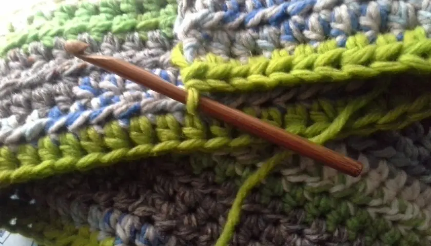 Coperta ai ferri con avanzi di lana, un'opera in corso con un uncinetto di legno che lavora maglie verdi su uno sfondo multicolore.