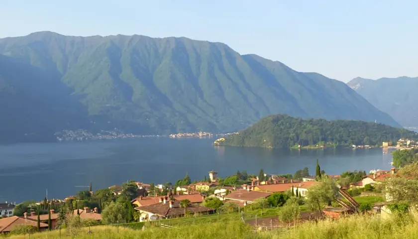 Villaggio sulle rive del lago più grande d'Europa, con montagne verdi sullo sfondo e un prato fiorito in primo piano.