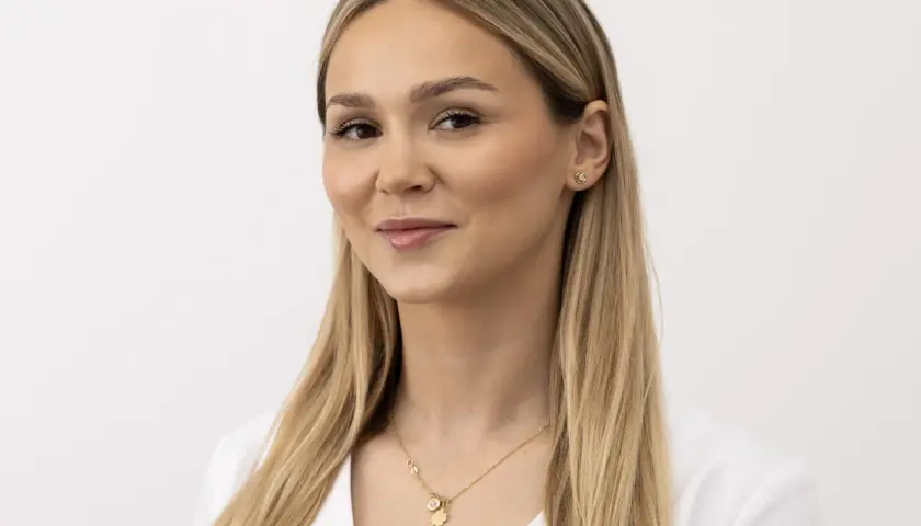 Kobieta z blond włosami i delikatnym makijażem, uśmiecha się lekko, choć jej nienaturalny wyraz twarzy sugeruje, że pozuje do zdjęcia.