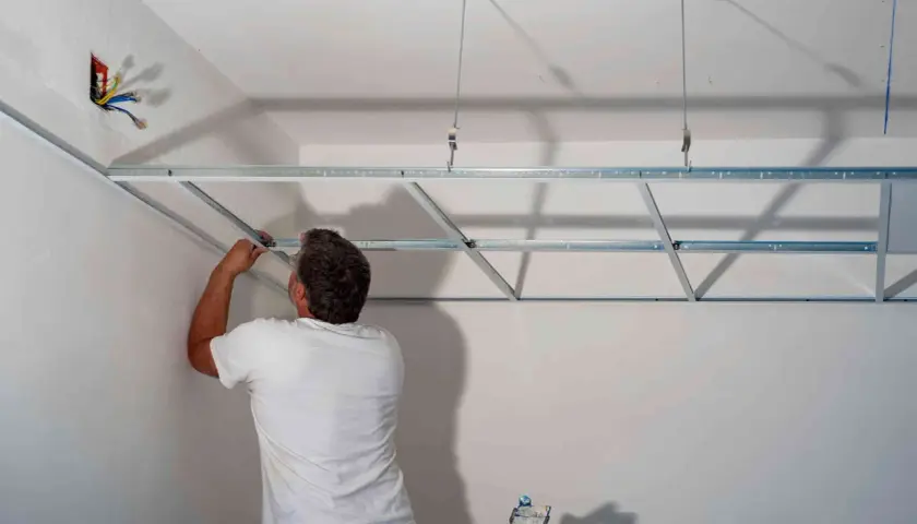 Mężczyzna w białej koszulce montuje metalowy stelaż pod sufity podwieszane. Widać przewody elektryczne w ścianie.