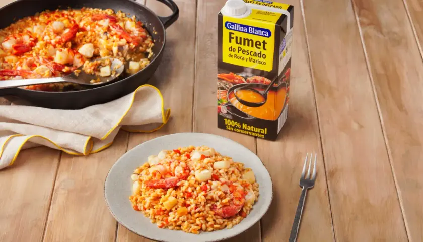 Arroz meloso con gambas y trozos de pescado en paellera y plato, junto a un cartón de fumet.