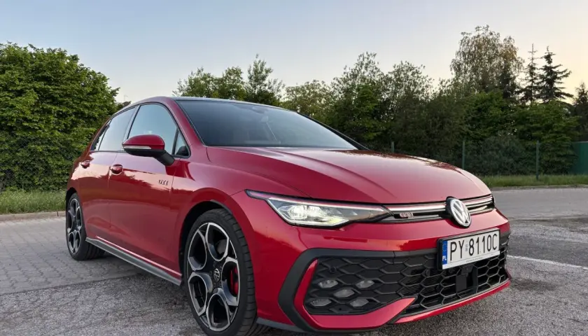 Czerwony Volkswagen Golf GTI na parkingu, z zielenią w tle.