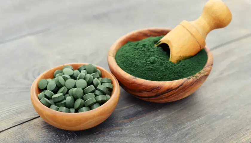 Due ciotole in legno: una con compresse verdi e l'altra con polvere di **pianta alga spirulina**, con un cucchiaino di legno.