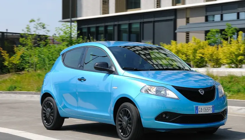 Lancia Ypsilon 1.3 Multijet Diesel azzurra in movimento, con sfondo di edifici moderni e vegetazione.