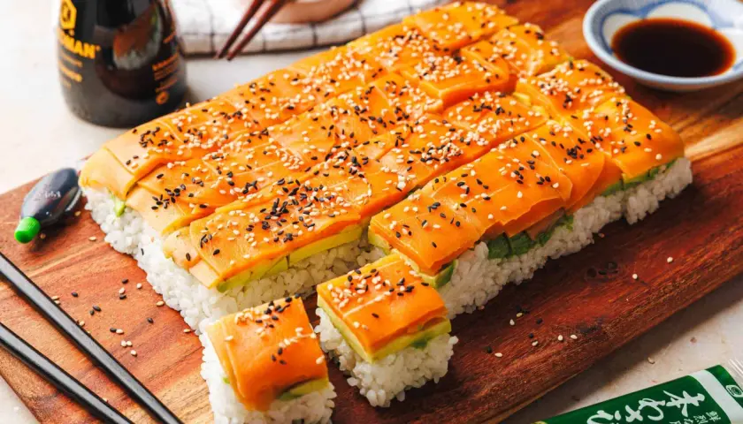 Rechteckiges Sushi mit Reis, Avocado und orangefarbener veganer Sushi Füllung, bestreut mit Sesam. Sojasauce und Wasabi sind daneben.
