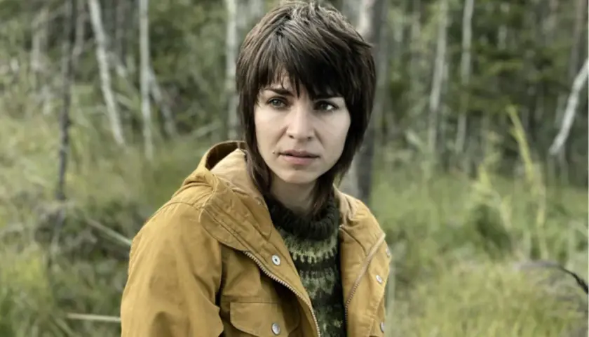 Ida Elise Broch i en gul jacka och gröntröja, med en skog i bakgrunden.