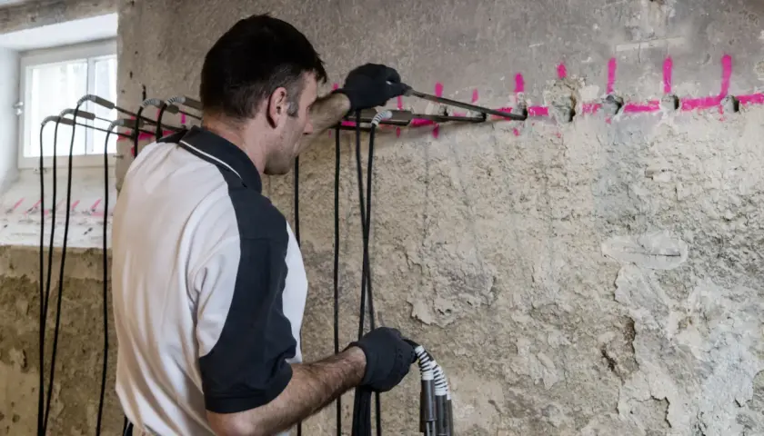 Mann führt eine Horizontalsperre im Mauerwerk durch. Bohrlöcher mit pinker Markierung und Kabel sind sichtbar.
