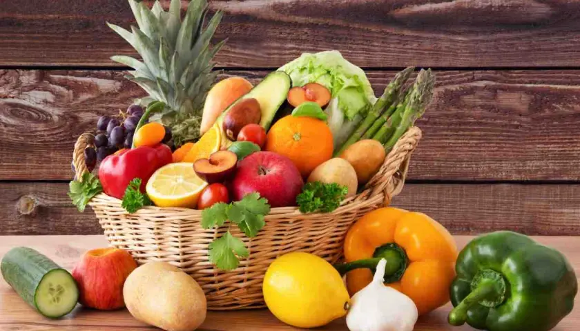 Cesto pieno di frutta e verdura fresca: ananas, uva, mele, arance, limoni, pomodori, peperoni, patate, cetrioli, asparagi e lattuga.