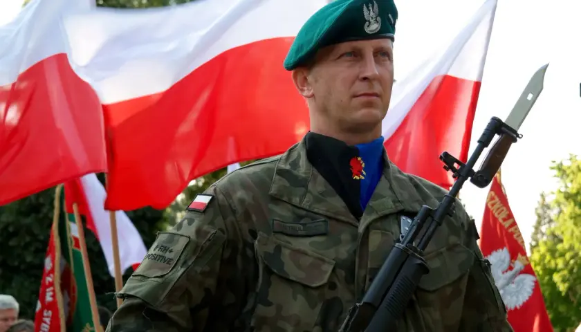 Żołnierz w zielonym beretcie, z karabinem i bagnetem, na tle dwóch biało-czerwonych flag.