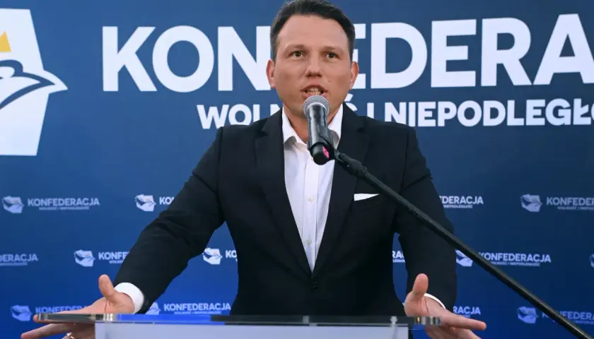 Mężczyzna w garniturze przemawia do mikrofonu na tle baneru Konfederacji. Dyskutuje, dlaczego KO chce odebrać dotacje partii opozycyjnej poprzez nowelizację kodeksu wyborczego.