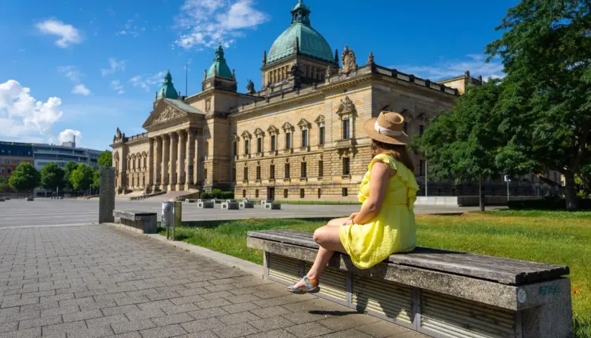 Kobieta w słomkowym kapeluszu i żółtej sukience siedzi na ławce przed monumentalnym budynkiem w Lipsku, mieście w Saksonii znanym z międzynarodowych targów.