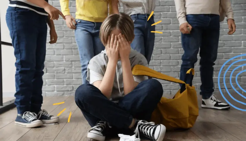Chłopiec siedzi na podłodze, zakrywając twarz dłońmi, podczas gdy inne dzieci wskazują na niego. To obraz tego, czym jest bullying.
