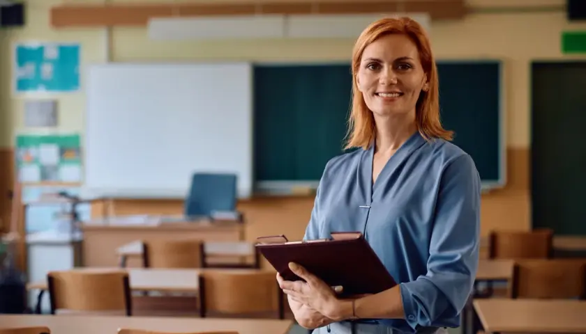 Uśmiechnięta nauczycielka z rudymi włosami trzyma teczkę w pustej sali lekcyjnej. Czas na zasłużony urlop nauczyciela!