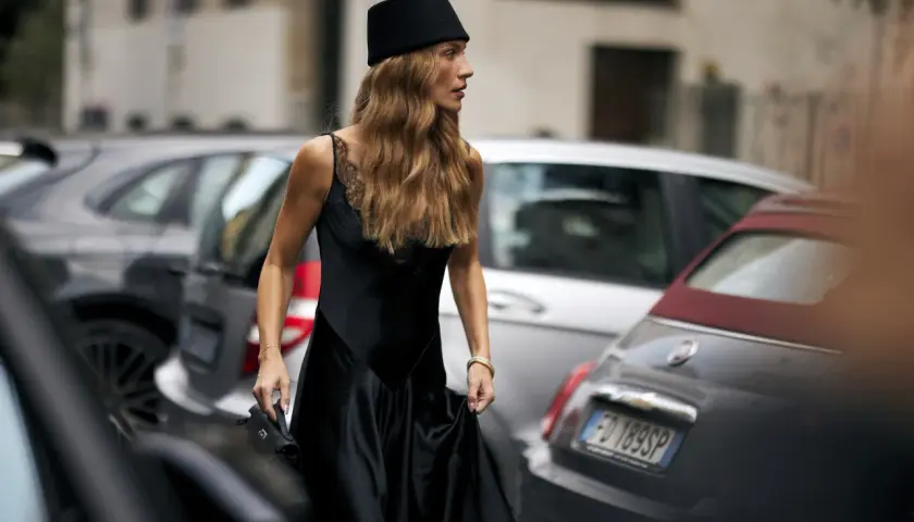Mujer con sombrero negro y vestido de seda, su pelo en pausa, camina entre coches aparcados.