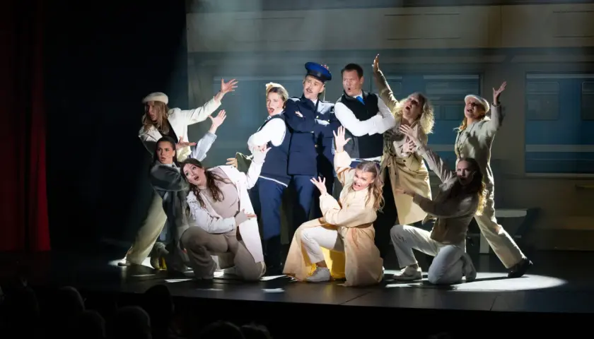 Ensemblen i en svensk revy, med en polisman i centrum, gestikulerar dramatiskt på scenen framför en tågvagnsbakgrund.