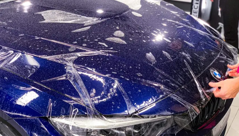 Nakładanie folii ochronnej na maskę ciemnoniebieskiego BMW. Widać krople wody i pęcherzyki powietrza pod folią.