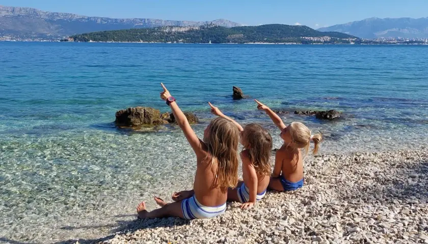 Trzy dziewczynki siedzą na kamienistej plaży w Chorwacji, wskazując palcami na morze. Idealne miejsce, gdzie do Chorwacji z dziećmi.