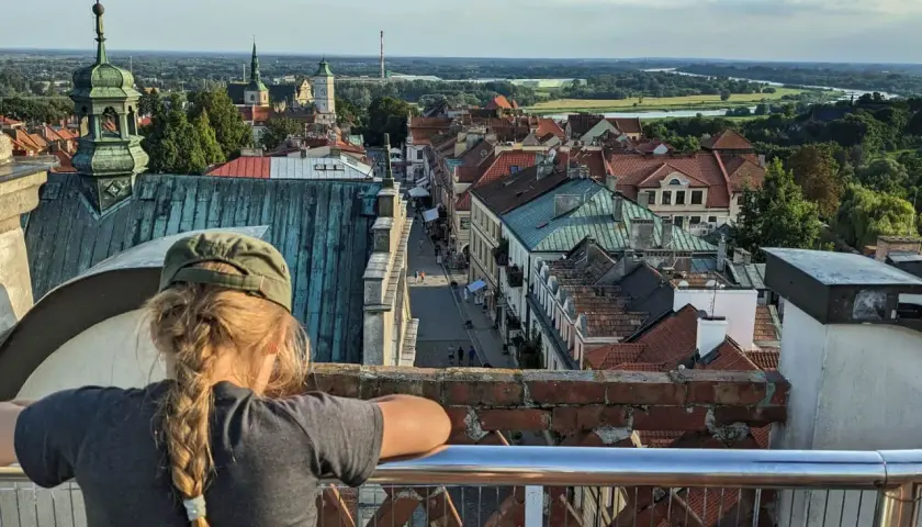 Widok z góry na Sandomierz, z dziewczynką w czapce na pierwszym planie. W oddali widać Wisłę i charakterystyczną Brama Opatowska.
