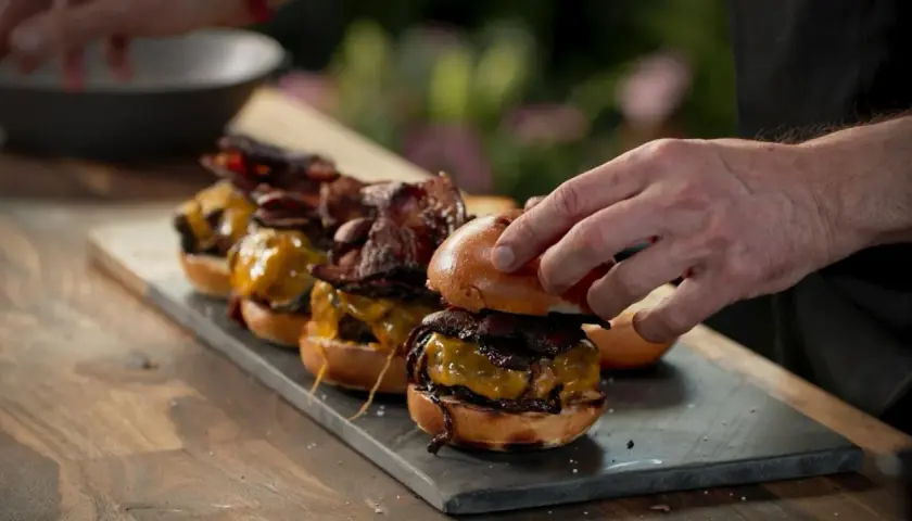 En hand lägger på toppen av en smashed burger med ost och bacon. Flera likadana burgare står uppradade.
