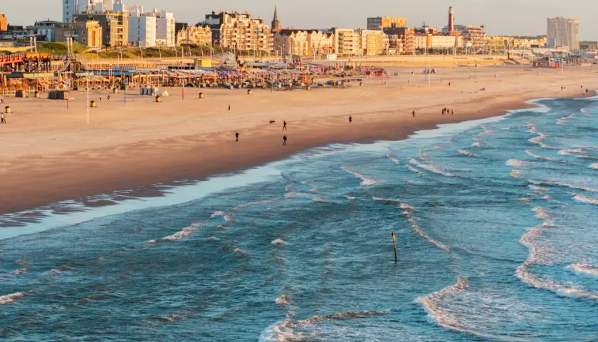 Ein sonniger Tag an der Nordsee Holland. Wellen brechen am Strand, im Hintergrund die Stadtkulisse mit Hotels und Strandpavillons.