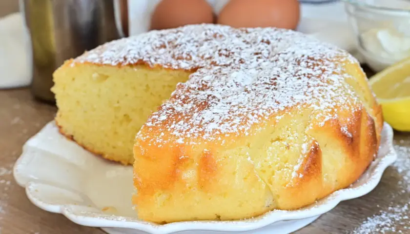 Una soffice torta al limone, spolverata di zucchero a velo, pronta per essere gustata. Ingredienti freschi come uova e limone sullo sfondo.